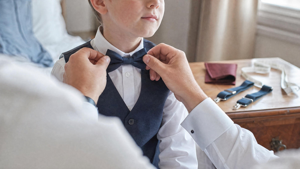 Un adulte ajuste un nœud papillon sur un enfant en chemise blanche, avec un mètre ruban visible sur une commode, dans une chambre lumineuse avant une cérémonie Image